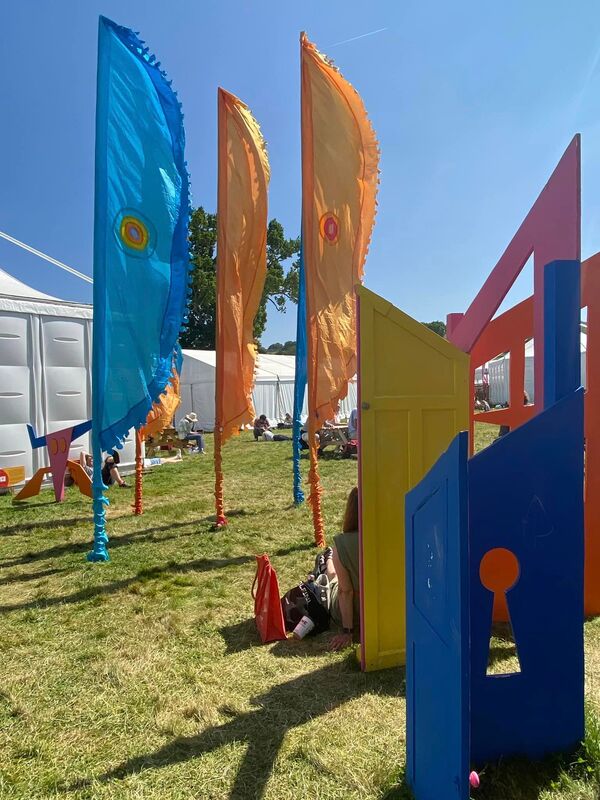 One of the real-world sculptures from the AR experience – it looks like a series of interlinked doors painted blue, pink, orange and yellow. Blue and orange Hay Festival flags are in the background.